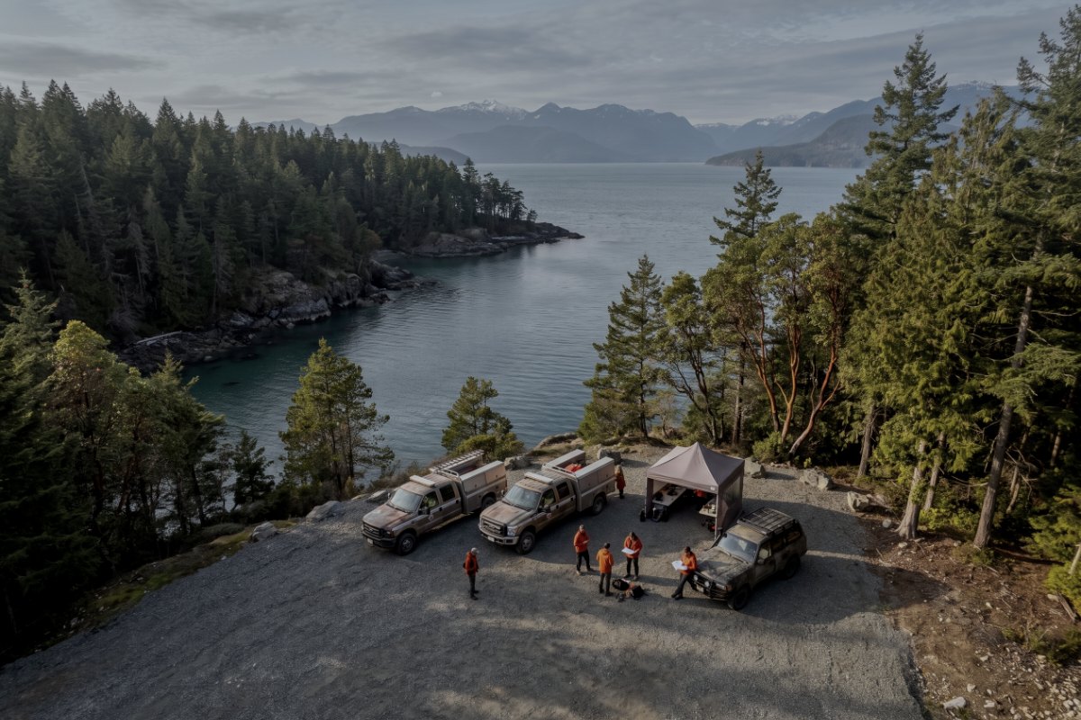 Aerial view of a search and rescue staging area on a forest road landing above an inlet, with trucks, a command tent and searchers