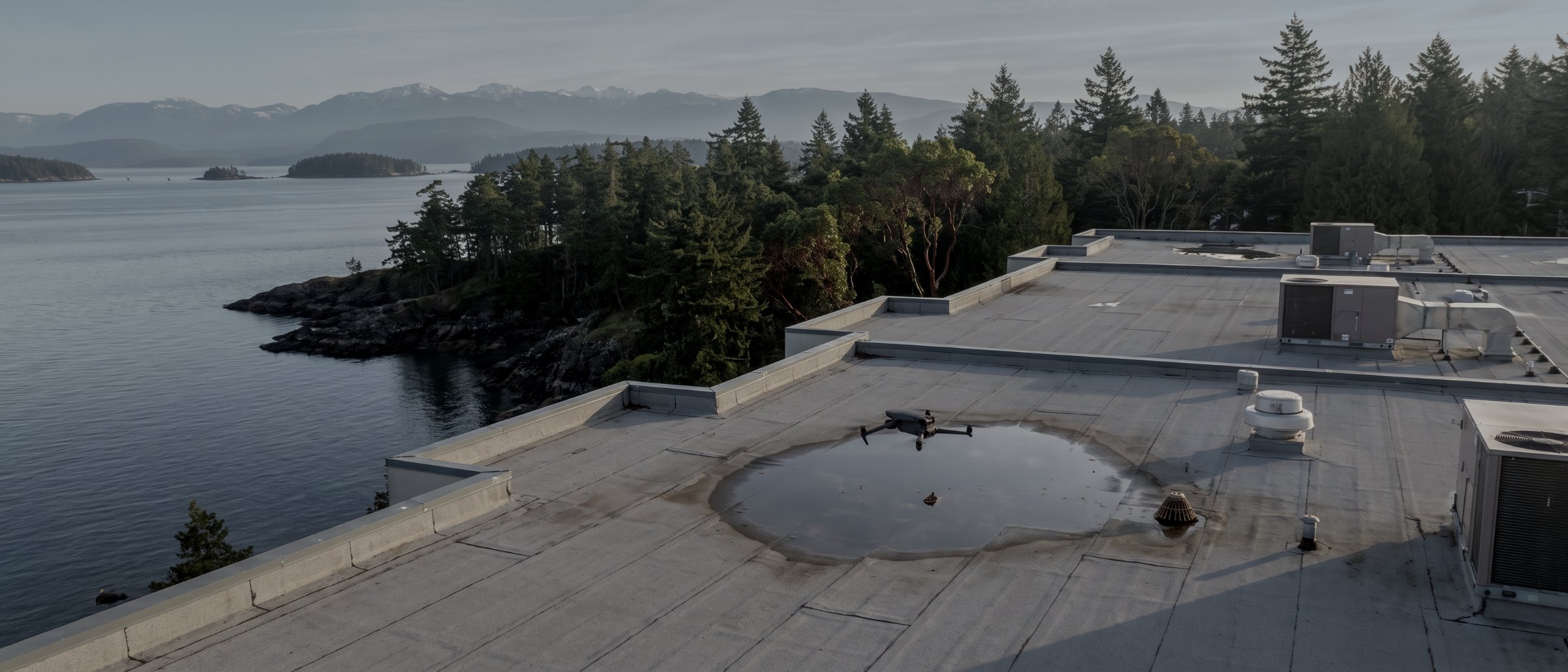 Aerial view of a flat commercial roof with HVAC units and a patch of ponded water, the coastline and forest beyond