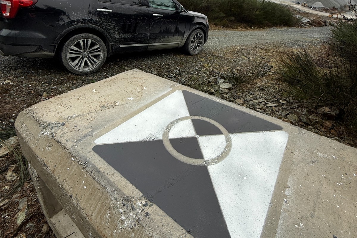A painted concrete survey control monument at a Vancouver Island quarry, its centre punch mark visible between black and white quadrants, with the Elite Drone Solutions vehicle and quarry stockpiles behind