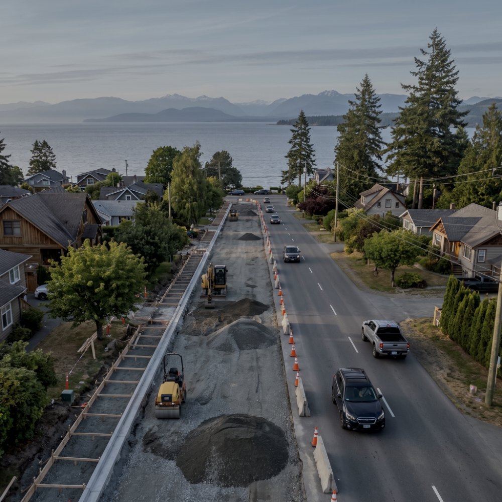 Aerial view of a road reconstruction in a coastal town, one lane excavated with a roller and cones while traffic passes