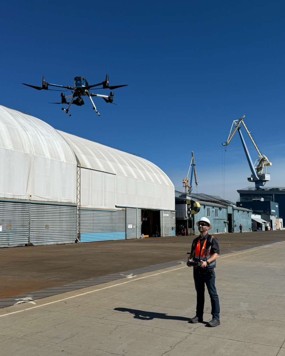 An Elite Drone Solutions pilot in hard hat and hi-vis at the controller on a working shipyard apron, the M350 RTK in the air overhead and gantry cranes behind